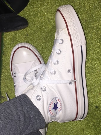 A pair of white high-top sneakers with a rubber sole and a red and blue logo on the side. The sneakers have white laces and silver eyelets. They are being worn by someone wearing grey pants and are photographed on a green textured surface.
