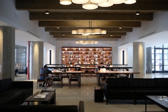 A modern library or study space featuring large wooden tables with people sitting and reading or working on laptops. The room has exposed wooden beams on the ceiling and is illuminated by hanging circular lights. A bookshelf filled with books and decorative items stands prominently against the far wall. The space is well-organized and conducive to quiet study or work.