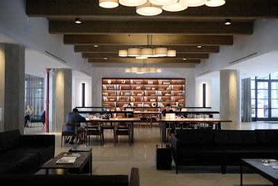 A modern library or study space featuring large wooden tables with people sitting and reading or working on laptops. The room has exposed wooden beams on the ceiling and is illuminated by hanging circular lights. A bookshelf filled with books and decorative items stands prominently against the far wall. The space is well-organized and conducive to quiet study or work.