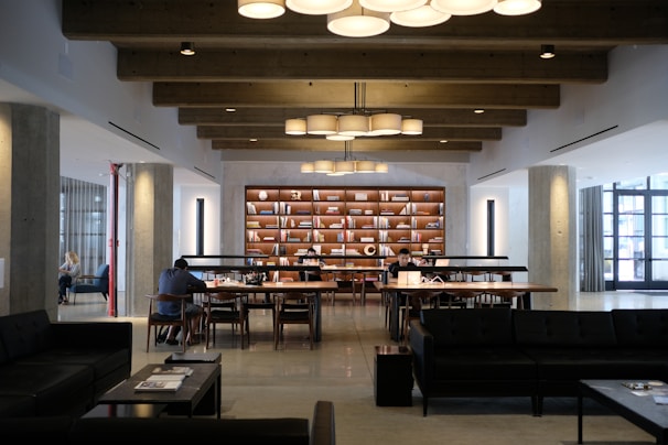 A modern library or study space featuring large wooden tables with people sitting and reading or working on laptops. The room has exposed wooden beams on the ceiling and is illuminated by hanging circular lights. A bookshelf filled with books and decorative items stands prominently against the far wall. The space is well-organized and conducive to quiet study or work.