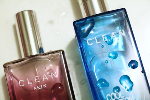 Two bottles of perfume are placed at an angle, one with a pink tint labeled 'Clean Skin' and the other with a blue tint labeled 'Clean Cool Cotton'. Both have reflective silver caps and droplets that mimic water, adding a refreshing look.