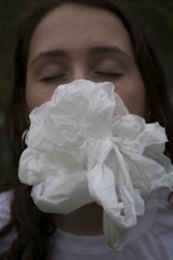 A person with their eyes closed is holding a crumpled white plastic bag or fabric against their face, obscuring their mouth and nose. The background is dark and out of focus.