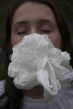 A person with their eyes closed is holding a crumpled white plastic bag or fabric against their face, obscuring their mouth and nose. The background is dark and out of focus.