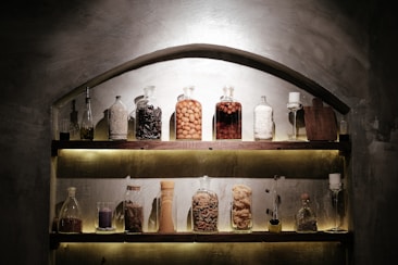 A rustic wooden shelf displays various glass jars and bottles filled with different ingredients, such as spices, pasta, and liquids. There are also small decorative objects like a purple candle and a wooden cutting board. Soft lighting enhances the warm, vintage atmosphere of the space.