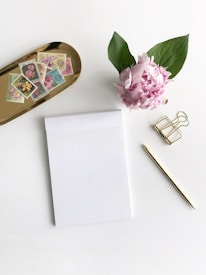 A neat arrangement on a white surface includes a small stack of floral-themed postage stamps on a gold tray, a single pink peony with green leaves, a blank notepad, a gold pen, and a gold geometric paper clip.
