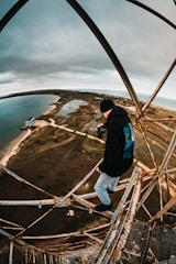 A person wearing a black beanie and holding a camera stands on an old, rusted metal structure high above the ground. The landscape below stretches into the distance, featuring a coastline with sandy beaches, bodies of water, and sparse vegetation under a cloudy sky.