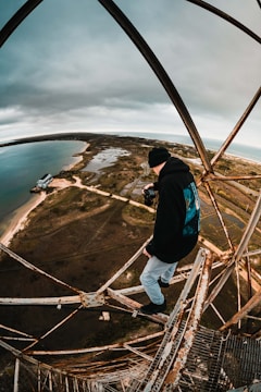 A person wearing a black beanie and holding a camera stands on an old, rusted metal structure high above the ground. The landscape below stretches into the distance, featuring a coastline with sandy beaches, bodies of water, and sparse vegetation under a cloudy sky.