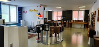 The room is a modern audio equipment showroom featuring various speakers and sound systems displayed on stands and shelves. The walls are decorated with colorful words like pop, hip-hop, and house, indicating different music genres. The space is well-lit with large windows allowing in natural light, and the floor is polished. Several tables hold additional audio equipment, and there are soundproofing panels on one of the walls.