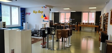 The room is a modern audio equipment showroom featuring various speakers and sound systems displayed on stands and shelves. The walls are decorated with colorful words like pop, hip-hop, and house, indicating different music genres. The space is well-lit with large windows allowing in natural light, and the floor is polished. Several tables hold additional audio equipment, and there are soundproofing panels on one of the walls.
