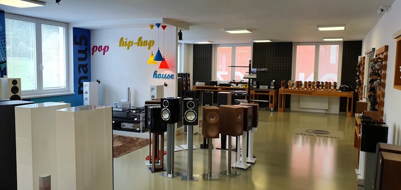 The room is a modern audio equipment showroom featuring various speakers and sound systems displayed on stands and shelves. The walls are decorated with colorful words like pop, hip-hop, and house, indicating different music genres. The space is well-lit with large windows allowing in natural light, and the floor is polished. Several tables hold additional audio equipment, and there are soundproofing panels on one of the walls.