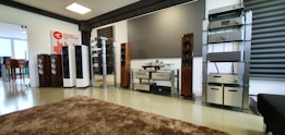 A spacious audio equipment showroom featuring a variety of high-end sound systems. Several tall speakers are aligned against the walls, displaying sleek wooden and metallic finishes. Electronics such as amplifiers and receivers are arranged on modern glass shelves, and a large, plush brown rug covers the floor. A sign on the left side suggests a focus on luxury electronics.