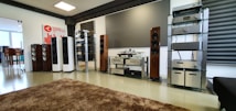 A spacious audio equipment showroom featuring a variety of high-end sound systems. Several tall speakers are aligned against the walls, displaying sleek wooden and metallic finishes. Electronics such as amplifiers and receivers are arranged on modern glass shelves, and a large, plush brown rug covers the floor. A sign on the left side suggests a focus on luxury electronics.