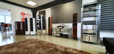 A spacious audio equipment showroom featuring a variety of high-end sound systems. Several tall speakers are aligned against the walls, displaying sleek wooden and metallic finishes. Electronics such as amplifiers and receivers are arranged on modern glass shelves, and a large, plush brown rug covers the floor. A sign on the left side suggests a focus on luxury electronics.