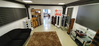 The room is elegantly designed with a variety of modern audio equipment. There are multiple loudspeakers of different sizes positioned around the room, a large sofa, and a brown rug on the floor. On the right side, there is a glass shelf with audio components including a turntable. The walls are adorned with large acoustic panels and there's a display with a logo reading 'Luksusmi Elektronika'. The room is well-lit and has a contemporary, high-tech vibe.