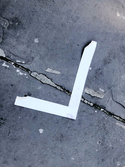 A white tape arranged in an L-shape on a cracked concrete surface. The concrete has several visible cracks and a rough texture, with a mix of grey shades and small spots of dirt.