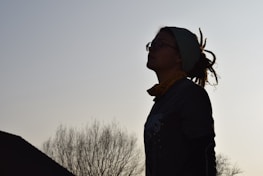 A silhouetted person with dreadlocks and glasses is looking up towards the sky. They are outdoors, with bare tree branches visible against a clear sky.