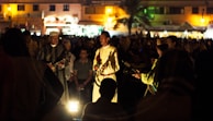 A group of musicians is performing in a dimly lit outdoor setting. They are surrounded by a crowd of people, some sitting and others standing. The musicians wear traditional attire and are playing string instruments, while a soft light illuminates them from below, creating a warm ambiance.