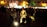 A group of musicians is performing in a dimly lit outdoor setting. They are surrounded by a crowd of people, some sitting and others standing. The musicians wear traditional attire and are playing string instruments, while a soft light illuminates them from below, creating a warm ambiance.