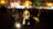 A group of musicians is performing in a dimly lit outdoor setting. They are surrounded by a crowd of people, some sitting and others standing. The musicians wear traditional attire and are playing string instruments, while a soft light illuminates them from below, creating a warm ambiance.