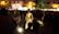 A group of musicians is performing in a dimly lit outdoor setting. They are surrounded by a crowd of people, some sitting and others standing. The musicians wear traditional attire and are playing string instruments, while a soft light illuminates them from below, creating a warm ambiance.