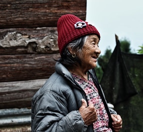 An elderly person with a warm smile is seen in profile, wearing a red knitted hat adorned with flower patterns and a thick grey jacket. The background features wooden logs and some greenery, giving a rustic and natural atmosphere.