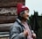 An elderly person with a warm smile is seen in profile, wearing a red knitted hat adorned with flower patterns and a thick grey jacket. The background features wooden logs and some greenery, giving a rustic and natural atmosphere.