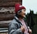 An elderly person with a warm smile is seen in profile, wearing a red knitted hat adorned with flower patterns and a thick grey jacket. The background features wooden logs and some greenery, giving a rustic and natural atmosphere.