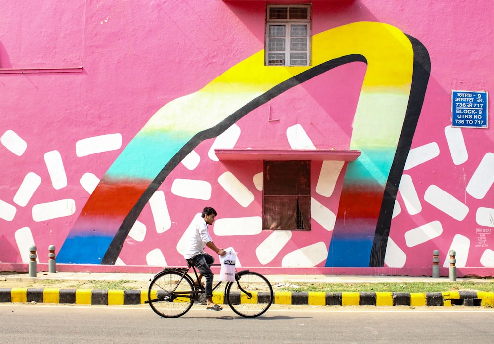 man riding bicycle along pink building