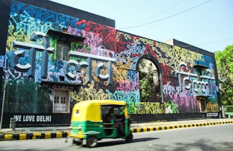 A vibrant wall mural with colorful graffiti art covers a large building facade. The graffiti is composed of various colors and abstract designs with prominent text in a bold, three-dimensional style. A green and yellow auto rickshaw is moving on the road in front of the mural. There are trees visible in the background and the phrase 'We Love Delhi' is written on the building.