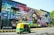 A vibrant wall mural with colorful graffiti art covers a large building facade. The graffiti is composed of various colors and abstract designs with prominent text in a bold, three-dimensional style. A green and yellow auto rickshaw is moving on the road in front of the mural. There are trees visible in the background and the phrase 'We Love Delhi' is written on the building.