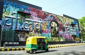 A vibrant wall mural with colorful graffiti art covers a large building facade. The graffiti is composed of various colors and abstract designs with prominent text in a bold, three-dimensional style. A green and yellow auto rickshaw is moving on the road in front of the mural. There are trees visible in the background and the phrase 'We Love Delhi' is written on the building.