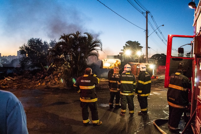 A group of firefighters wearing uniforms with reflective stripes stand near a fire truck at the scene of a fire. Smoke rises in the background as they seem to be coordinating their efforts under streetlights and dusk sky. The scene includes debris and a construction vehicle nearby, suggesting cleanup or containment efforts.