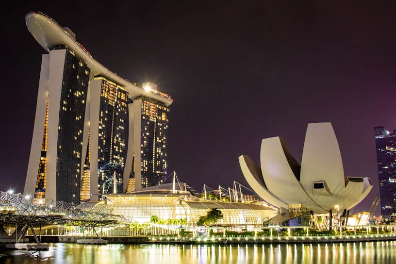 A nighttime cityscape featuring a large, illuminated hotel with a unique design resembling a ship on top of three towers. Nearby, there is a building shaped like a lotus flower, also brightly lit. The reflections of these structures are visible in the calm water in the foreground.