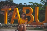 A person is sitting in front of large yellow letters that spell TABU. The individual is wearing a yellow shirt and is smiling. The background features greenery and some red-tinted branches.