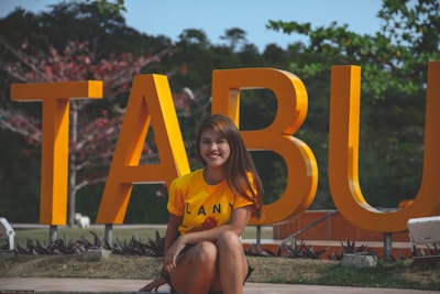 A person is sitting in front of large yellow letters that spell TABU. The individual is wearing a yellow shirt and is smiling. The background features greenery and some red-tinted branches.