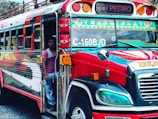 A brightly colored bus with the label 'Gabriela' is parked, with a man standing at the entrance. The bus has vibrant red, green, and yellow decorations with multiple lights on the front. The destination sign reads 'San Pedro'.