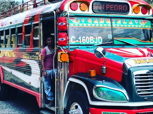 A brightly colored bus with the label 'Gabriela' is parked, with a man standing at the entrance. The bus has vibrant red, green, and yellow decorations with multiple lights on the front. The destination sign reads 'San Pedro'.