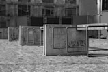 A series of concrete barriers adorned with graffiti stand in an open area. The ground appears dry and rough, and the background features a partially visible, decrepit building facade.