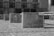 A series of concrete barriers adorned with graffiti stand in an open area. The ground appears dry and rough, and the background features a partially visible, decrepit building facade.