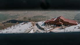 A snowy mountain landscape with a brown building featuring a red roof is prominently situated on the slope. Ski lifts and small cabins are visible, with a vast expanse of mountainous terrain in the background.