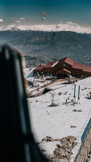 A ski resort with a prominent wooden lodge sits amid snow-covered grounds. Beyond the lodge, a mountain range is visible under a clear sky, while some ski lifts stand idle in the foreground.