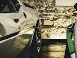 A close-up view of a polished white car parked indoors with a rough stone wall in the background. The lighting casts a warm glow, highlighting the car's sleek design and shiny wheels.