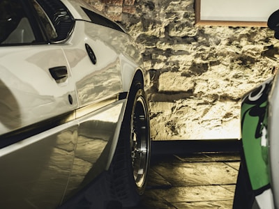 A close-up view of a polished white car parked indoors with a rough stone wall in the background. The lighting casts a warm glow, highlighting the car's sleek design and shiny wheels.