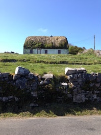 A quaint thatched-roof cottage sits atop a small grassy hill, surrounded by a rustic stone wall. The cottage has a white exterior with red window and door frames. The thatch appears weathered yet intact, with hints of green moss. A clear blue sky stretches out above, while a single utility pole with wires is visible to the right.