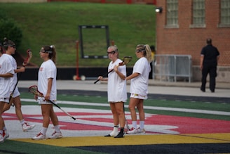 Several women in athletic gear are standing on an outdoor sports field holding lacrosse sticks. They are wearing white uniforms with visible team logos and appear to be engaged in a practice session or a game. The field has distinct colored lines and markings, and there's a brick building in the background.