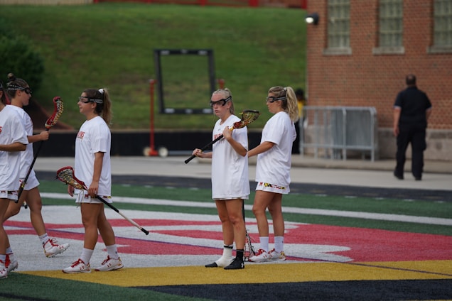 Several women in athletic gear are standing on an outdoor sports field holding lacrosse sticks. They are wearing white uniforms with visible team logos and appear to be engaged in a practice session or a game. The field has distinct colored lines and markings, and there's a brick building in the background.