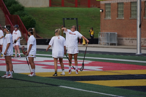 Several women are gathered on a sports field, wearing athletic gear, and holding lacrosse sticks. They appear to be in a casual setting, possibly during a practice or warm-up. The background features a building with brick walls and steps leading up to a grass area.