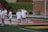 Several women are gathered on a sports field, wearing athletic gear, and holding lacrosse sticks. They appear to be in a casual setting, possibly during a practice or warm-up. The background features a building with brick walls and steps leading up to a grass area.