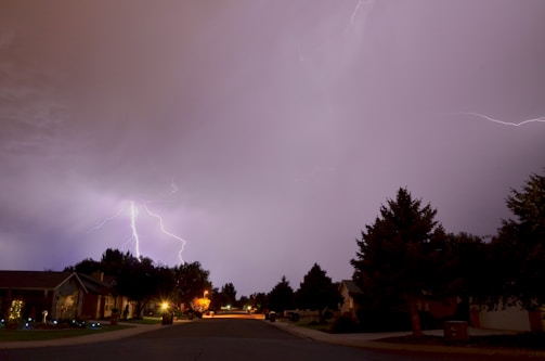 A residential street is illuminated by a bright lightning strike in the distance. Trees line the sidewalks, and houses with lit windows are visible on either side of the street. The sky is overcast, with some clouds reflecting the light from the lightning.