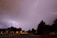 A residential street is illuminated by a bright lightning strike in the distance. Trees line the sidewalks, and houses with lit windows are visible on either side of the street. The sky is overcast, with some clouds reflecting the light from the lightning.